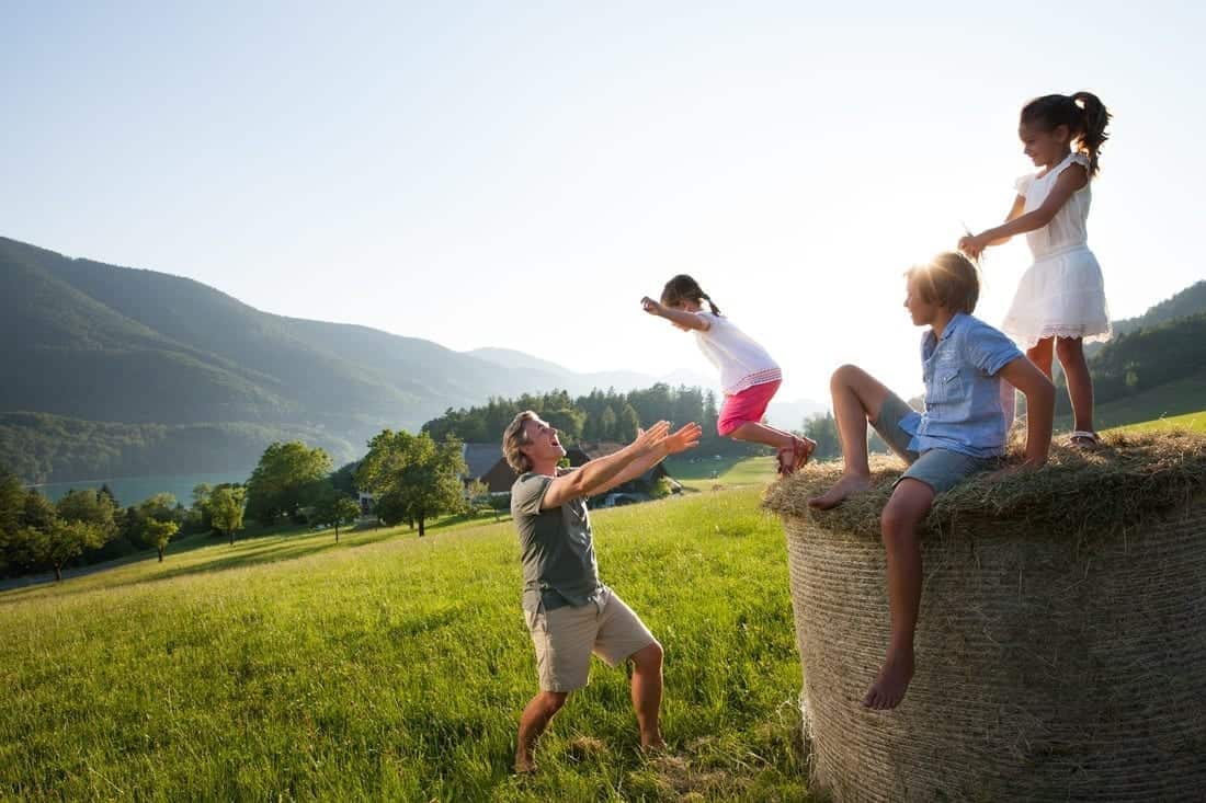 Familienzeit im Freien: Kinder springen von einer Heuballen auf eine Wiese mit Bergblick.