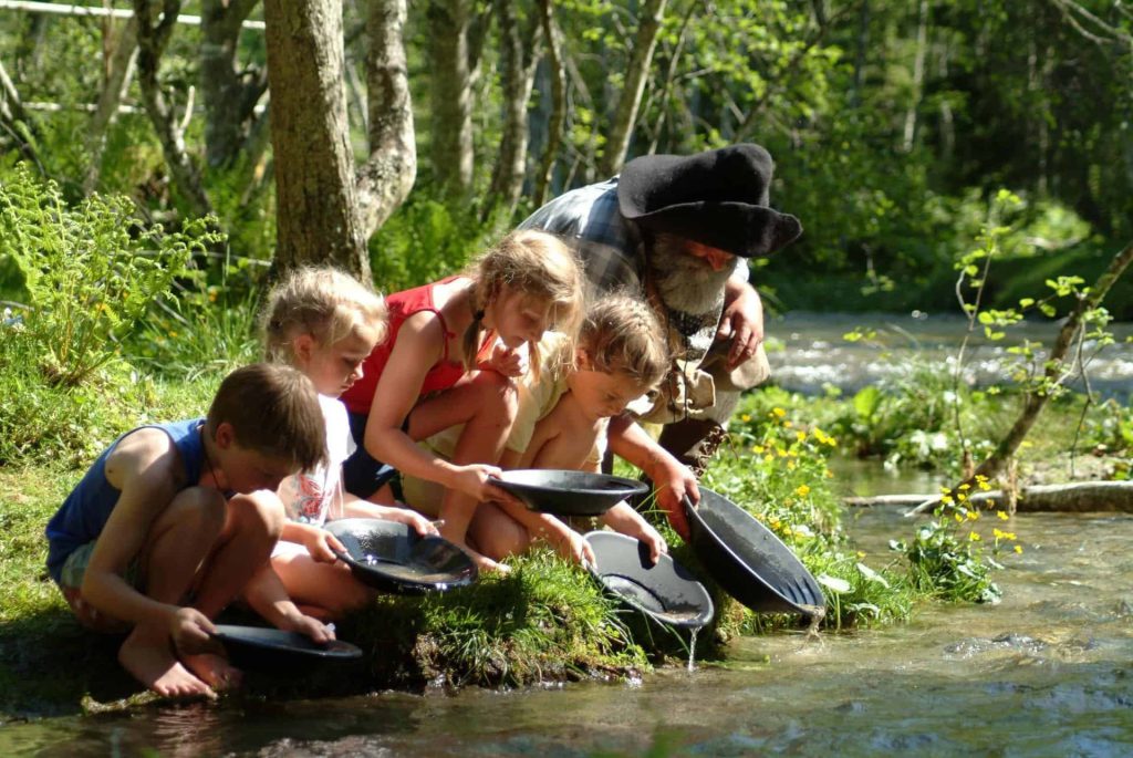 Children and an adult searching for gold by the river, surrounded by greenery and nature.