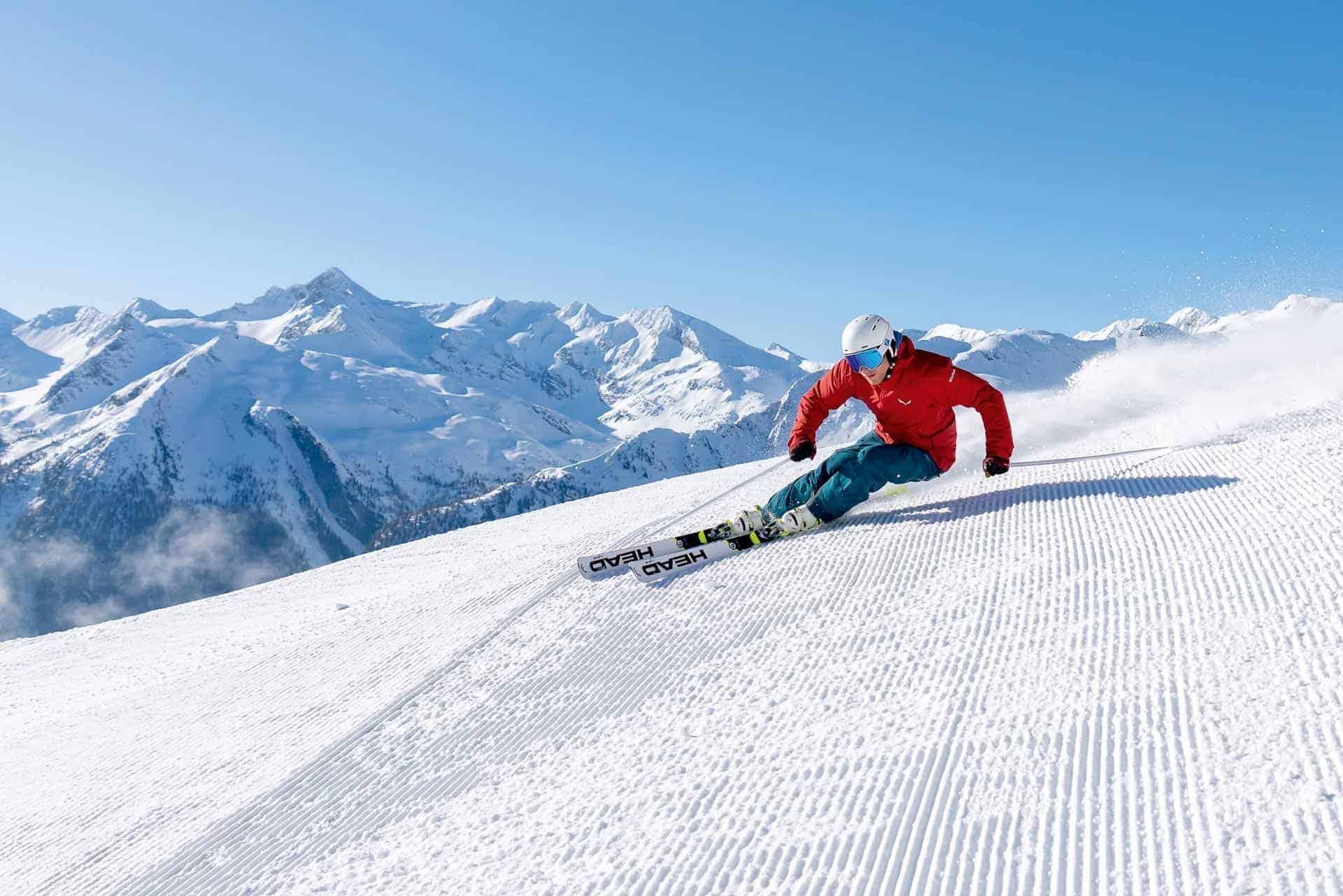Skifahrer auf einer Piste mit schneebedeckten Bergen im Hintergrund, klare Himmel.