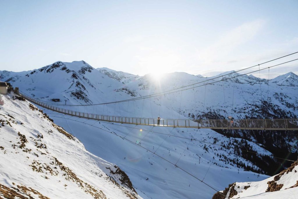 Schneebedeckte Berge mit einer Hängebrücke und Sonne im Hintergrund, ideal für Winterabenteuer.