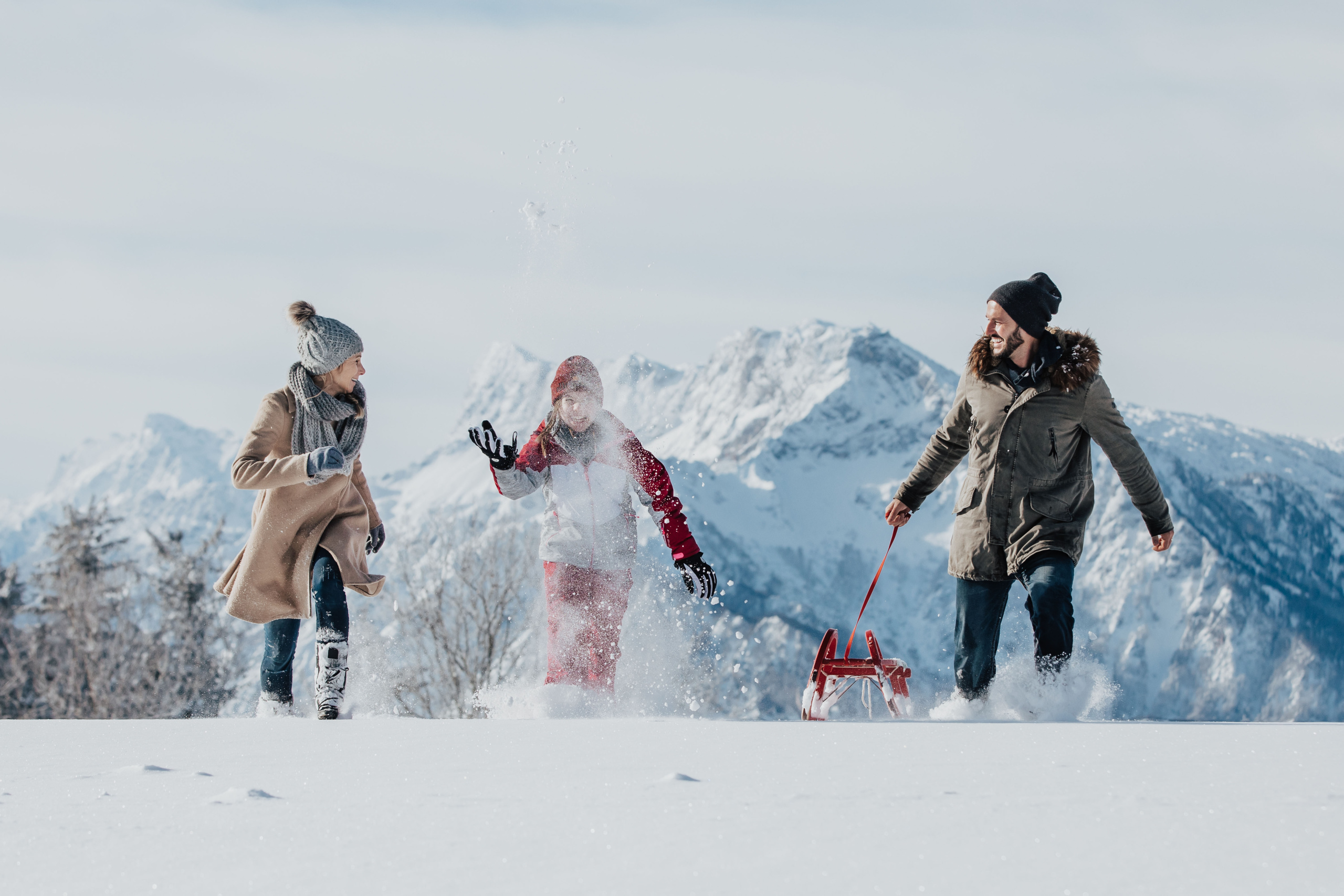 Schnee Family spielt im tiefen Schnee vor Bergen; rechts zieht eine Person einen roten Schlitten