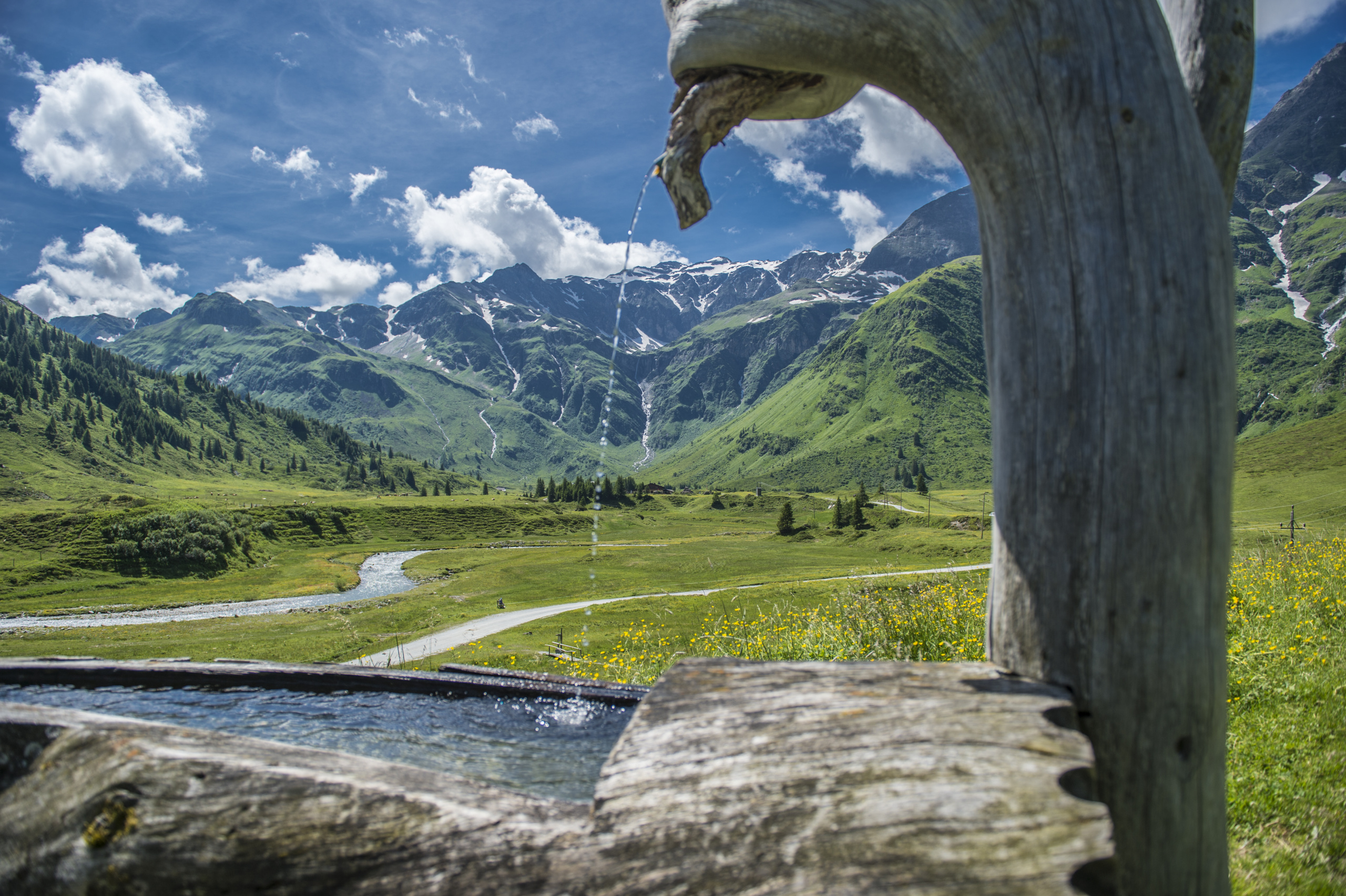 Holzbrunnen vor grüner Almwiese und schneebedeckten Bergen in Sportgastein