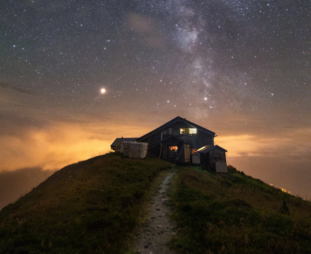 Berghütte auf einem Grashügel unter sternenklarem Himmel mit sichtbarer Milchstraße