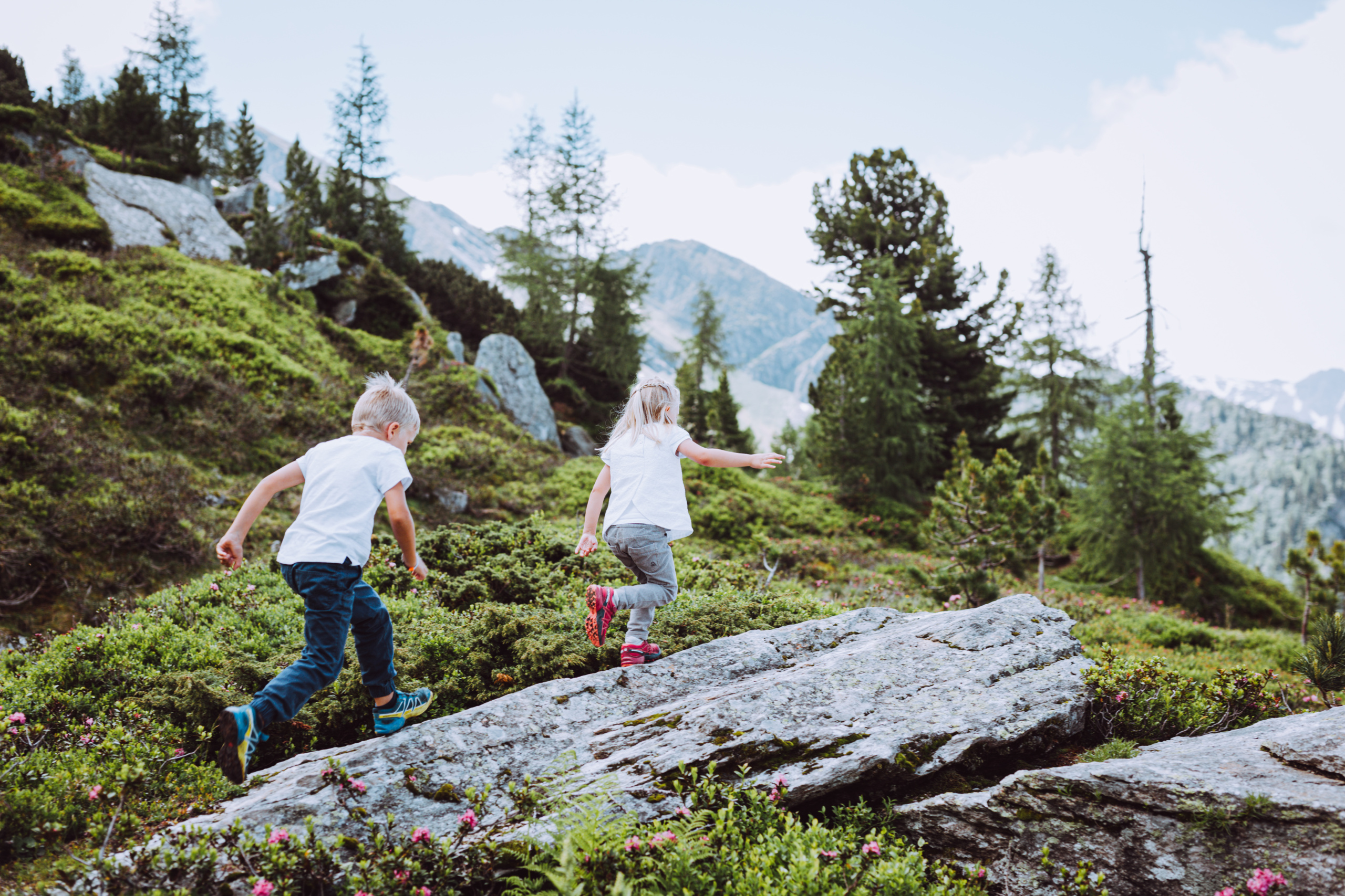 Zwei Kinder laufen über Felsen und Moos in einer alpinen Landschaft mit Lärchen und Bergen